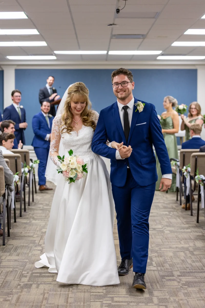 A bride in a white gown and a groom in a blue suit walk hand-in-hand down an aisle filled with seated guests in a decorated venue.