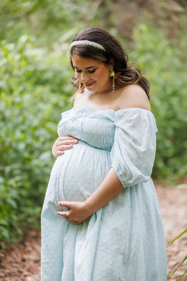 A pregnant woman in a light blue off-the-shoulder dress stands in a lush green forest, gently cradling her baby bump.