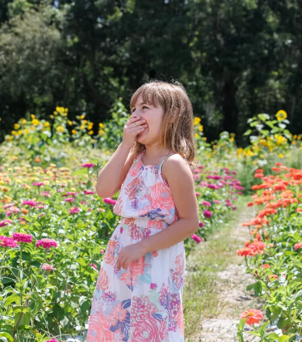 A young girl stands amidst vibrant flowers, wearing a colorful dress and touching her cheek, enjoying a sunny day in a garden.