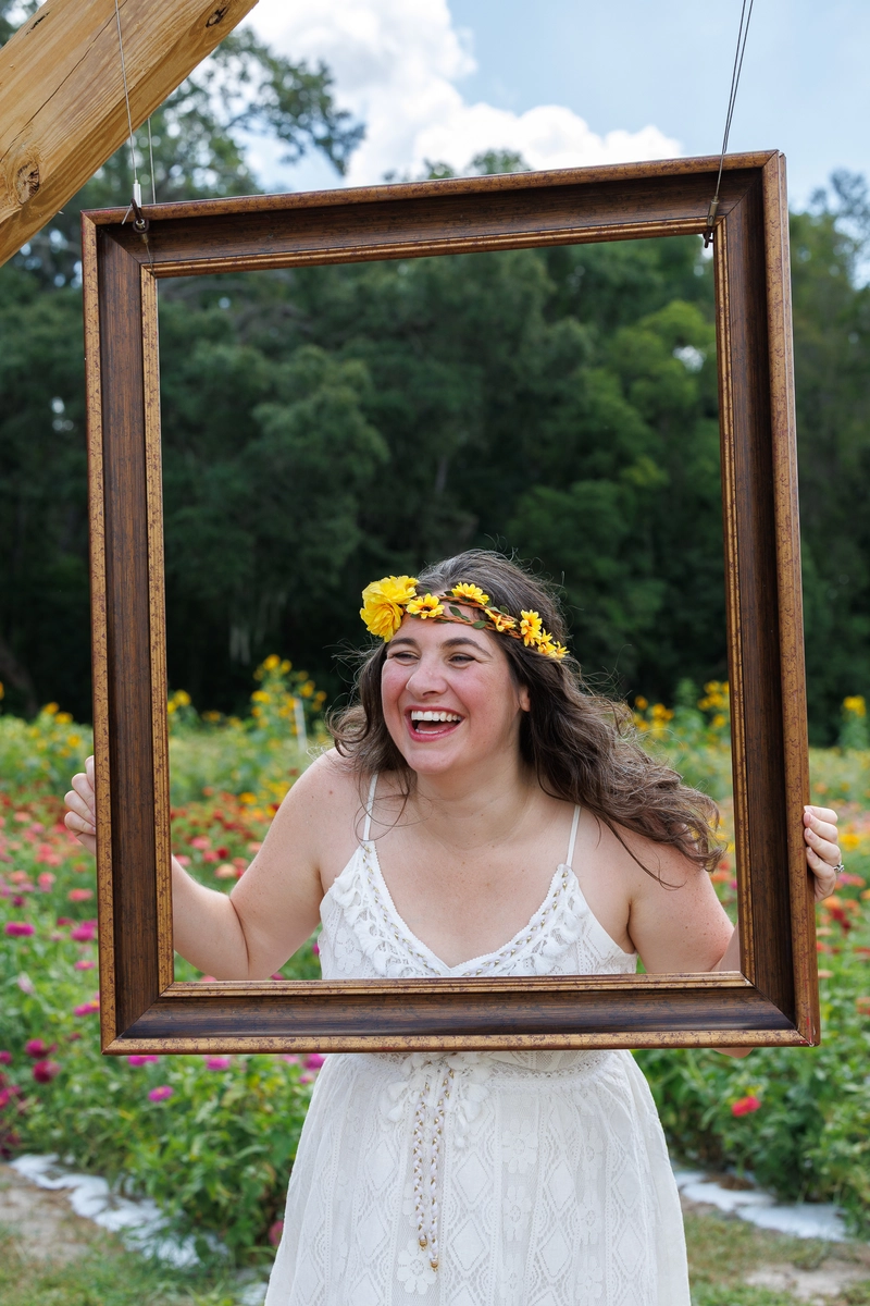 A woman in a white sundress holds a rustic wooden frame surrounded by a vibrant flower field under a blue sky.