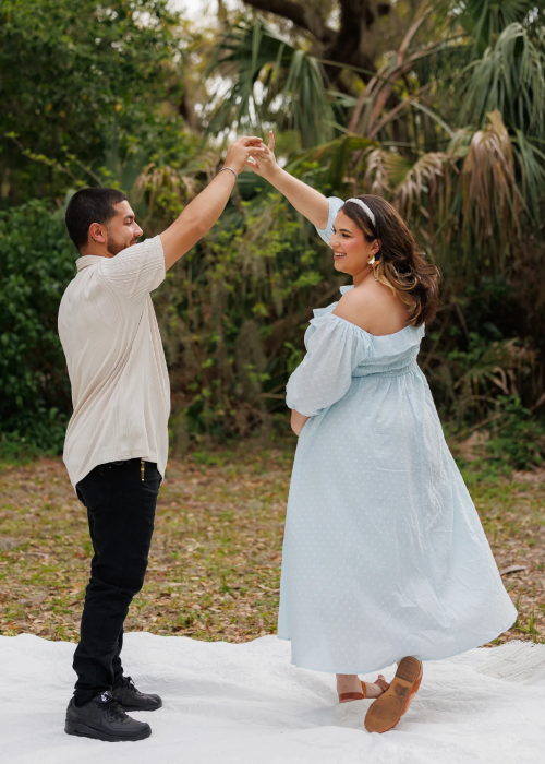 A couple joyfully dances together outdoors, surrounded by lush greenery, with the woman in a light blue dress and the man in a casual outfit