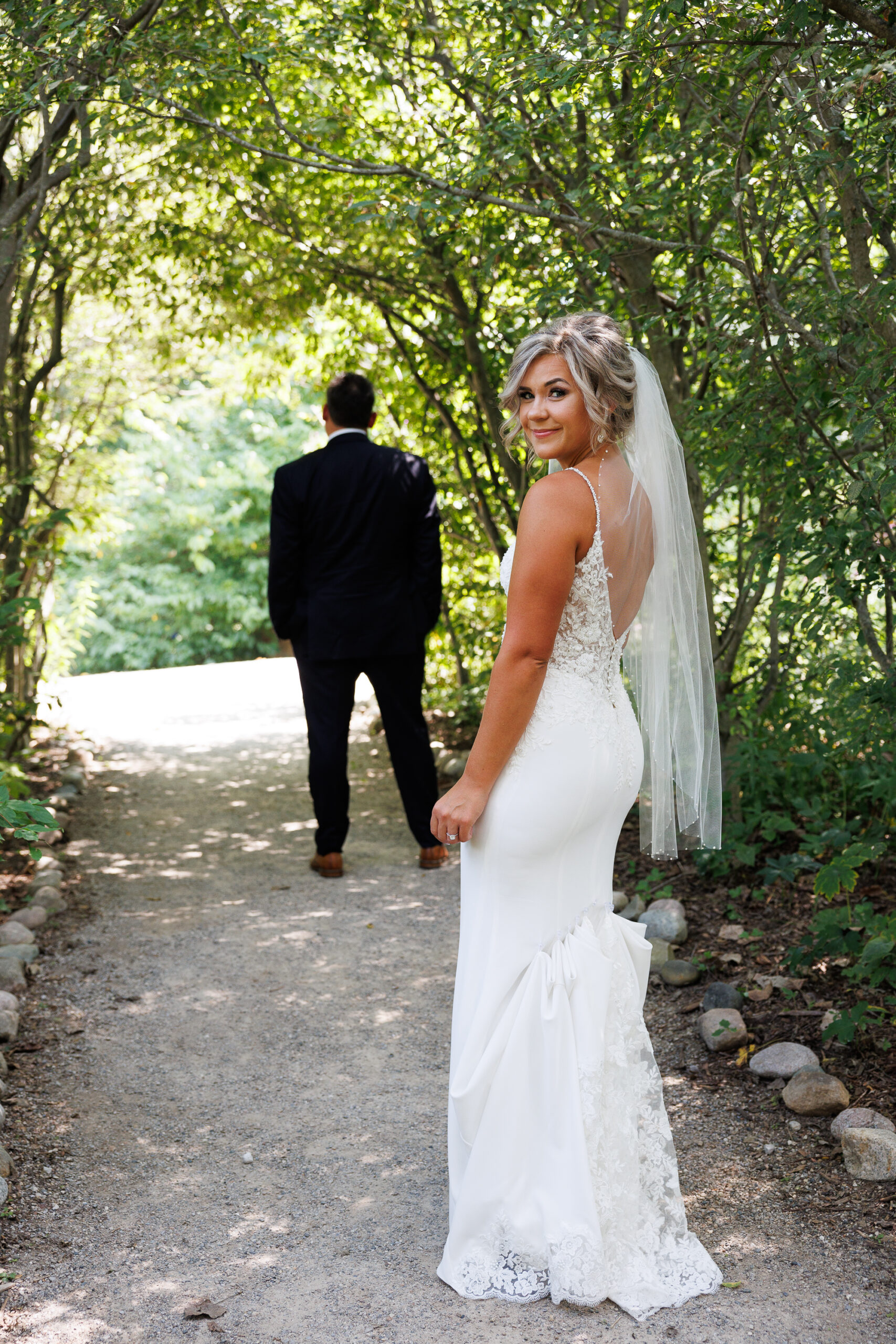 7N4A7440 A pregnant woman in a white dress stands among vibrant flowers, cradling her belly in a sunny, blooming garden.